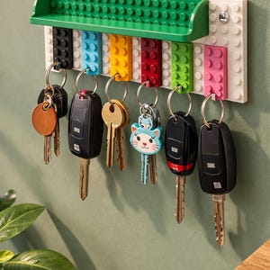 May include: A wall-mounted key holder constructed from colorful toy bricks. The key rack features a green shelf and a series of colored blocks, each holding a set of keys. The keys have various keychains, including a cat design.