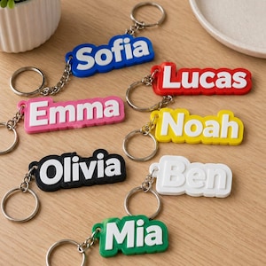 Puede incluir: Una colección de llaveros con nombres coloridos. Cada llavero presenta un nombre diferente en letras blancas sobre un fondo de color. Los llaveros están unidos a llaveros y cadenas plateadas. Los nombres incluyen Sofia, Lucas, Emma, Noah, Olivia, Ben y Mia.