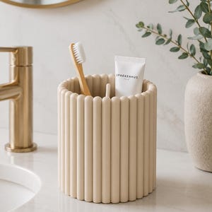 May include: A cylindrical beige bathroom organizer featuring a vertical ribbed texture on the exterior. The container holds a wooden toothbrush with white bristles and a white tube of Sprekenhus Oslo toothpaste, with a central divider inside.
