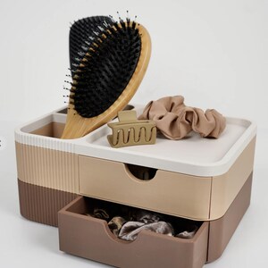 Puede incluir: Un organizador de varios compartimentos en tonos marr&oacute;n, beige y blanco, que contiene un cepillo para el pelo de madera, una pinza para el pelo beige y una goma para el pelo beige. El organizador tiene dos cajones y una bandeja superior.