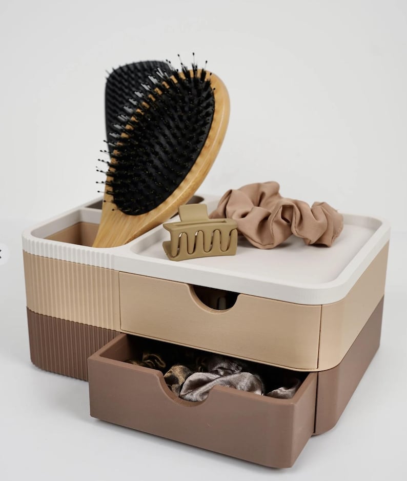Puede incluir: Un organizador de varios compartimentos en tonos marr&oacute;n, beige y blanco, que contiene un cepillo para el pelo de madera, una pinza para el pelo beige y una goma para el pelo beige. El organizador tiene dos cajones y una bandeja superior.