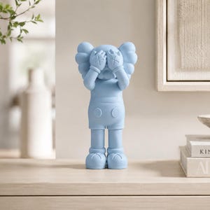 Puede incluir: Figura azul claro de un personaje de dibujos animados con los ojos tachados, cubri&eacute;ndose la cara con las manos. La figura est&aacute; sobre una superficie clara, con un jarr&oacute;n de vegetaci&oacute;n y libros al fondo. Mide unos 25 cm de altura.