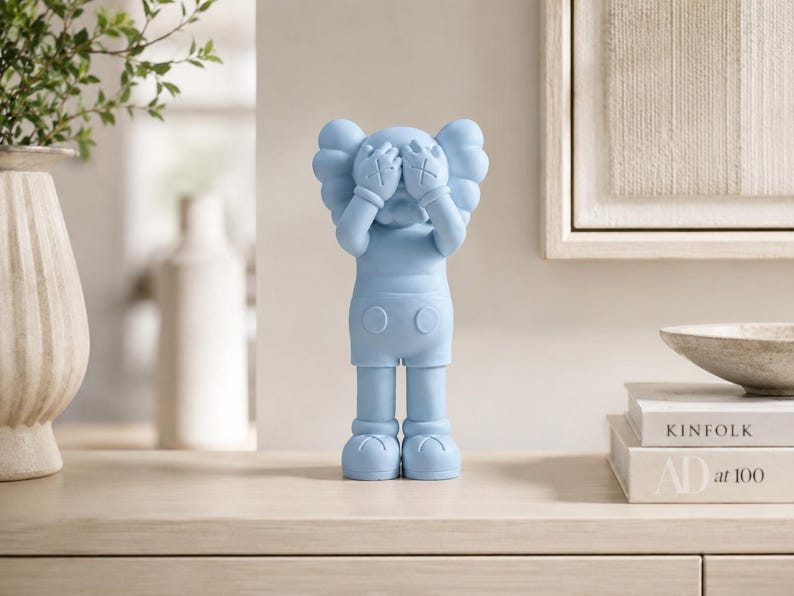 Puede incluir: Figura azul claro de un personaje de dibujos animados con los ojos tachados, cubri&eacute;ndose la cara con las manos. La figura est&aacute; sobre una superficie clara, con un jarr&oacute;n de vegetaci&oacute;n y libros al fondo. Mide unos 25 cm de altura.