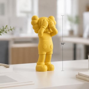 Puede incluir: Figura amarilla de un personaje de dibujos animados, de aproximadamente 25 cm de altura, con las manos cubriendo los ojos. La figura tiene una cabeza redondeada, pantalones cortos y pies grandes. La figura est&aacute; sobre una superficie blanca.