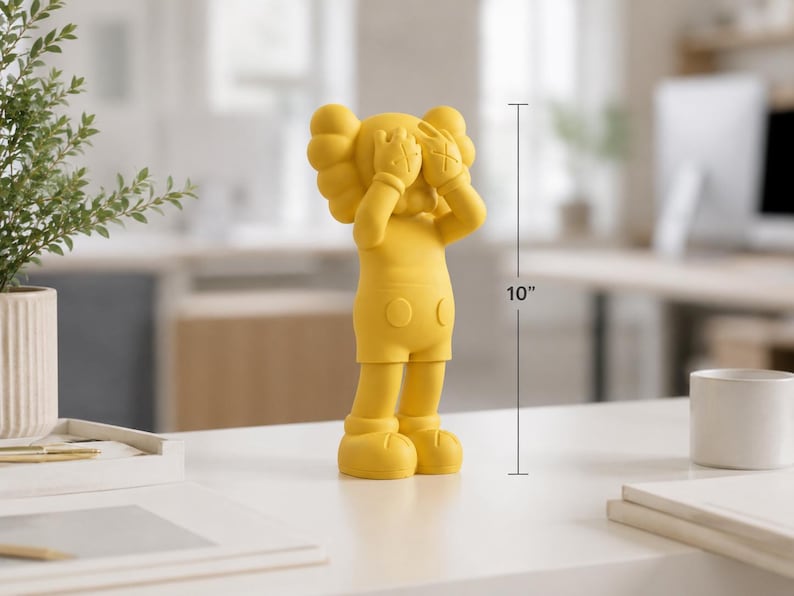 Puede incluir: Figura amarilla de un personaje de dibujos animados, de aproximadamente 25 cm de altura, con las manos cubriendo los ojos. La figura tiene una cabeza redondeada, pantalones cortos y pies grandes. La figura est&aacute; sobre una superficie blanca.