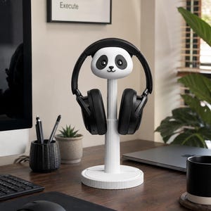 May include: A white headphone stand featuring a stylized panda head at the top, supported by a vertical ribbed column and a circular base. A pair of black over-ear headphones is resting on the stand, with the headband arched over the panda head.