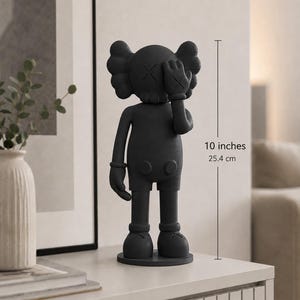 Puede incluir: Una figura gris oscuro de un personaje de dibujos animados, de aproximadamente 25,4 cm de altura. La figura tiene una cabeza redondeada con ojos en forma de X, una mano enguantada que cubre un ojo y pies redondeados. Se apoya sobre una base circular.