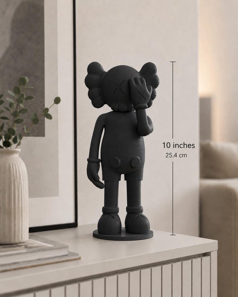 Puede incluir: Una figura gris oscuro de un personaje de dibujos animados, de aproximadamente 25,4 cm de altura. La figura tiene una cabeza redondeada con ojos en forma de X, una mano enguantada que cubre un ojo y pies redondeados. Se apoya sobre una base circular.