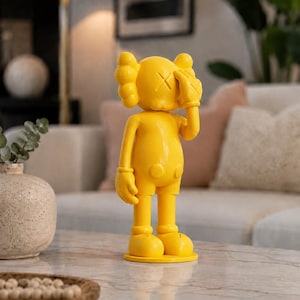 Puede incluir: Una figura de color amarillo brillante con un dise&ntilde;o &uacute;nico. La figura tiene una cabeza redonda con una "X" en cada ojo y una mano que cubre un ojo. La figura est&aacute; de pie sobre una base redonda. Es un objeto decorativo.