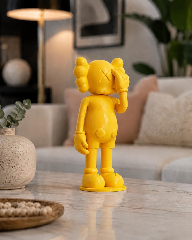 Puede incluir: Una figura de color amarillo brillante con un dise&ntilde;o &uacute;nico. La figura tiene una cabeza redonda con una "X" en cada ojo y una mano que cubre un ojo. La figura est&aacute; de pie sobre una base redonda. Es un objeto decorativo.