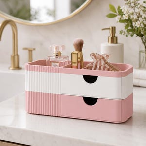Puede incluir: Un organizador de ba&ntilde;o rosa y blanco de dos niveles con un dise&ntilde;o acanalado. La secci&oacute;n superior contiene un frasco de perfume, un pincel de maquillaje y una pinza para el pelo. La secci&oacute;n inferior tiene un caj&oacute;n.