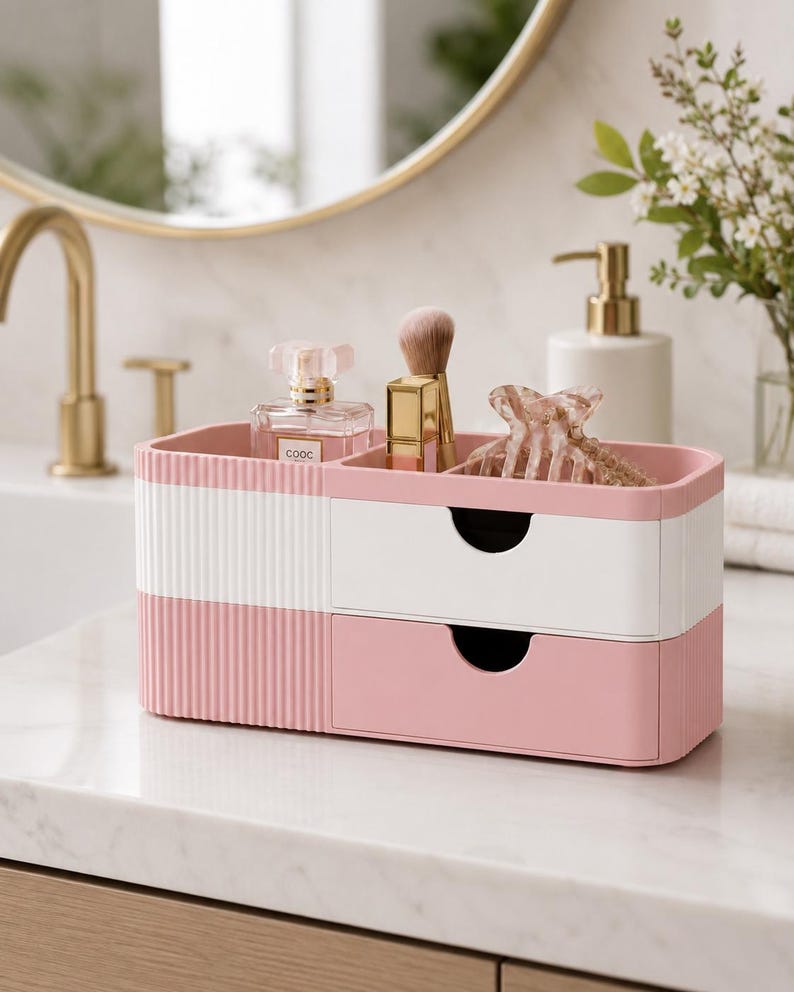 Puede incluir: Un organizador de ba&ntilde;o rosa y blanco de dos niveles con un dise&ntilde;o acanalado. La secci&oacute;n superior contiene un frasco de perfume, un pincel de maquillaje y una pinza para el pelo. La secci&oacute;n inferior tiene un caj&oacute;n.