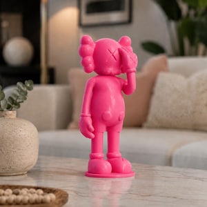 Puede incluir: Figura rosa brillante de un personaje de dibujos animados con ojos tachados y manos enguantadas cubriendo su rostro. La figura, sobre una base redonda, es un objeto de colecci&oacute;n de arte pop.