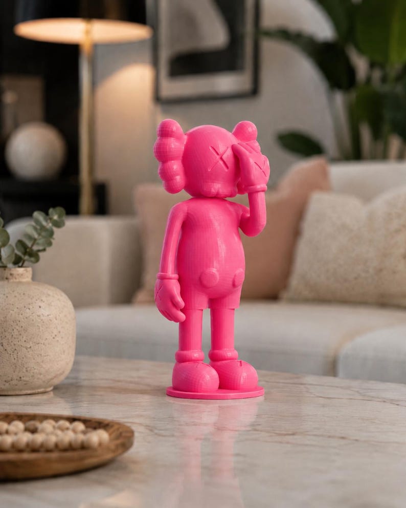 Puede incluir: Figura rosa brillante de un personaje de dibujos animados con ojos tachados y manos enguantadas cubriendo su rostro. La figura, sobre una base redonda, es un objeto de colecci&oacute;n de arte pop.