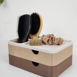 Puede incluir: Un organizador de dos cajones en tonos marr&oacute;n, beige y blanco, que contiene un cepillo para el cabello negro, un cepillo de madera, una pinza para el cabello beige y una goma para el pelo beige. El organizador est&aacute; dise&ntilde;ado para guardar accesorios de belleza.