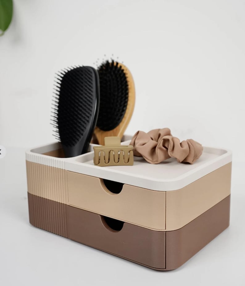Puede incluir: Un organizador de dos cajones en tonos marr&oacute;n, beige y blanco, que contiene un cepillo para el cabello negro, un cepillo de madera, una pinza para el cabello beige y una goma para el pelo beige. El organizador est&aacute; dise&ntilde;ado para guardar accesorios de belleza.