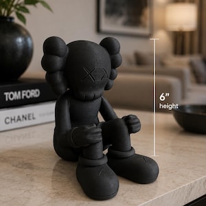Puede incluir: Una figura negra mate de un personaje sentado con orejas redondeadas, ojos en forma de X y manos enguantadas sobre las rodillas. La figura tiene una superficie texturizada y una indicación de altura de 15,24 cm junto al cuerpo.