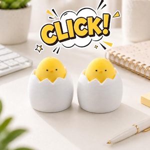 Pode incluir: Duas figuras de pintinhos amarelos saindo de metades de cascas de ovo brancas. Os pintinhos têm olhos pretos pequenos e estão colocados sobre uma superfície branca. Uma bolha de fala com a palavra "CLICK!" está acima dos pintinhos. A cena é em uma mesa com material de escritório.