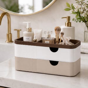 Puede incluir: Un organizador de dos niveles, marr&oacute;n, blanco y beige, con dos cajones y una bandeja superior. La bandeja contiene productos de belleza, incluyendo un frasco de perfume, brochas de maquillaje y productos para el cuidado de la piel. El organizador est&aacute; sobre una encimera de m&aacute;rmol.