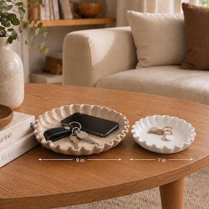May include: Two round ceramic trays with ruffled, scalloped edges. The larger tray is beige and measures 9 inches in diameter, holding a black smartphone and a set of keys. The smaller tray is white, measures 7 inches in diameter, and holds keys.