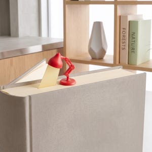 May include: A red desk lamp-shaped bookmark illuminates a page in a large, light gray book. The book is open, and the bookmark casts a yellow glow. The background includes a bookshelf with books and a decorative vase.