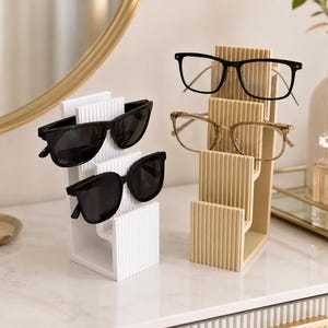 May include: Two vertical tiered eyewear stands, one white and one beige, each featuring a ribbed texture. The white stand holds two pairs of black sunglasses, while the beige stand holds one pair of black-rimmed glasses and one pair of clear-rimmed glasses.