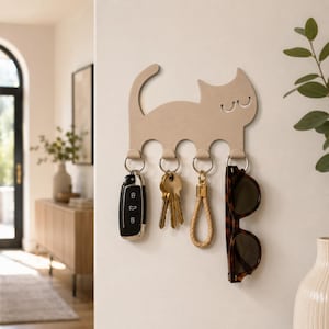 Pode incluir: Um suporte de parede bege para chaves em forma de gato com quatro ganchos de metal. Os ganchos seguram uma chave de carro preta, um molho de chaves, um porta-chaves de corda bege e óculos de sol castanhos em argolas prateadas.