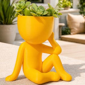 May include: A bright yellow planter in the shape of a person sitting with their arm resting on their head. The head of the figure is a bowl filled with green succulents. The planter is made of a smooth material and is designed for indoor or outdoor use.