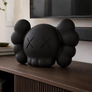 Könnte beinhalten: Eine mattschwarze KAWS Companion Totenkopffigur mit den charakteristischen X-förmigen Augen und abgerundeten, wolkenartigen Ohren. Die Skulptur hat eine glatte, solide Oberfläche mit einer stilisierten Ästhetik und einem breiten Kiefer.