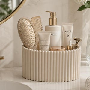 Può includere: Un organizer cilindrico color crema con superficie scanalata contenente vari prodotti di bellezza, tra cui una spazzola per capelli, un pettine, un flacone con erogatore marchiato OUAI, tubetti LAURA MERCIER e CAUDALIE, un contagocce e un rasoio dorato.