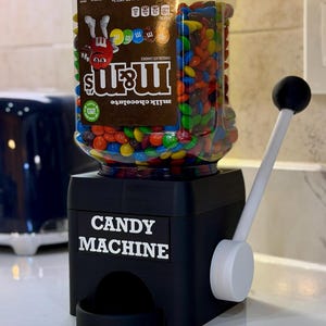 Könnte beinhalten: Ein schwarzer Süßigkeitenautomat aus Kunststoff mit weißem Hebel und einem durchsichtigen Kunststoffbehälter, gefüllt mit bunten M&M's Schokolinsen. Die Basis trägt die weiße Aufschrift CANDY MACHINE und der Behälter ist mit einem Etikett versehen.