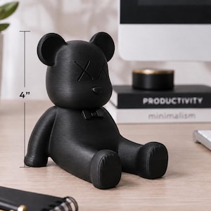 May include: A black bear figurine, approximately 4 inches tall, with crossed-out eyes and a bow tie. The bear is sitting with its legs extended. The figurine is on a wooden desk with a keyboard and books in the background.