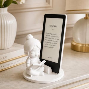 Pode incluir: Uma estatueta branca de uma menina lendo um livro, ao lado de um e-reader preto exibindo texto. O e-reader está apoiado em um suporte branco. O texto no e-reader diz "CHAPTER I".