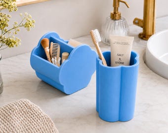 Puede incluir: Un juego de organizadores de baño azul claro con diseño de nube. Una pieza es un carrito con compartimentos que contiene una brocha de maquillaje, un peine y bastoncillos. La otra es un vaso cilíndrico con un cepillo de dientes y pasta dental.