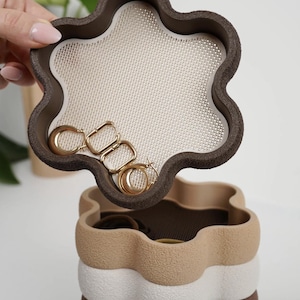 Peut inclure: Un organisateur de bijoux à plusieurs niveaux dans les tons de marron, beige et blanc. Le plateau supérieur, en forme de fleur, contient des boucles d'oreilles créoles dorées. L'organisateur a un design texturé et ondulé.