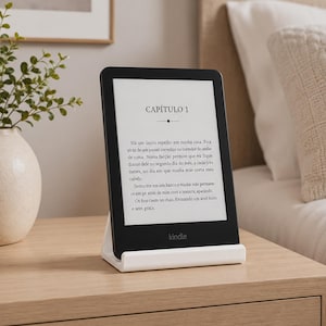 Op de afbeelding: Een zwarte Kindle e-reader met een wit scherm waarop Portugese tekst staat die begint met CAPÍTULO 1, rechtop geplaatst in een witte plastic standaard. Het apparaat heeft een dunne zwarte rand en het woord kindle onderaan in het midden.