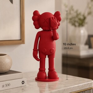 Puede incluir: Figura roja de un personaje de dibujos animados, de aproximadamente 25,4 cm de altura. La figura tiene una cabeza redonda con ojos en forma de X y una mano que cubre un ojo. Est&aacute; de pie sobre una base circular y lleva pantalones cortos.
