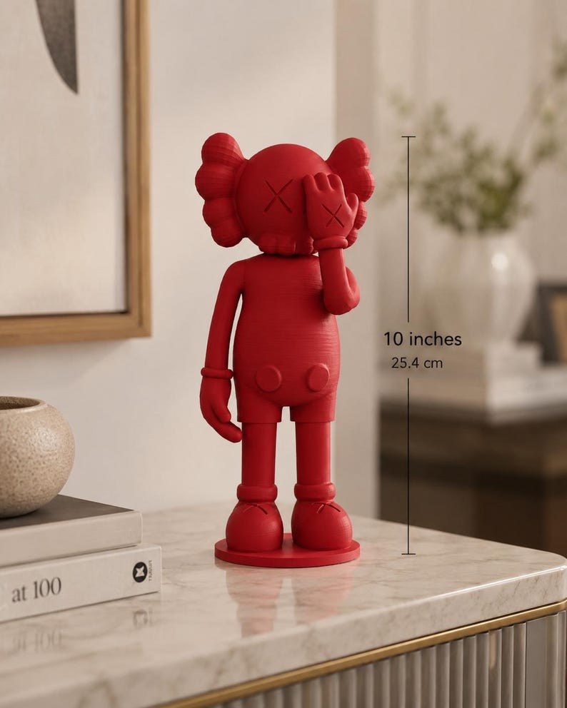 Puede incluir: Figura roja de un personaje de dibujos animados, de aproximadamente 25,4 cm de altura. La figura tiene una cabeza redonda con ojos en forma de X y una mano que cubre un ojo. Est&aacute; de pie sobre una base circular y lleva pantalones cortos.