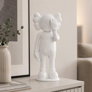 Puede incluir: Una figura blanca de un personaje de dibujos animados con los ojos tachados, cubri&eacute;ndose un ojo con una mano enguantada. La figura tiene una cabeza redonda con orejas estilizadas y est&aacute; de pie sobre una base circular. La figura est&aacute; sobre una superficie gris claro.