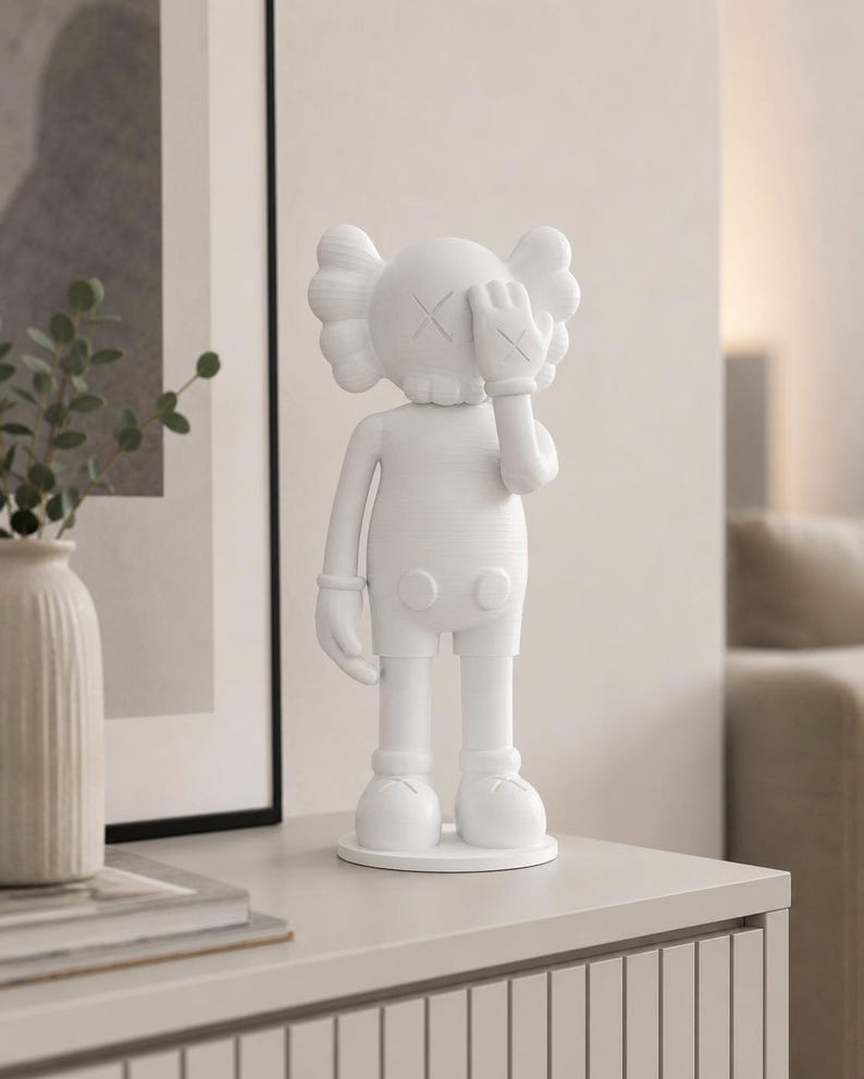 Puede incluir: Una figura blanca de un personaje de dibujos animados con los ojos tachados, cubri&eacute;ndose un ojo con una mano enguantada. La figura tiene una cabeza redonda con orejas estilizadas y est&aacute; de pie sobre una base circular. La figura est&aacute; sobre una superficie gris claro.
