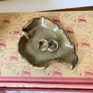 May include: A leaf-shaped ceramic trinket dish with a glossy, mottled olive-green glaze and dark brown edges. The dish features a small stem detail and holds a pair of small, silver-toned hoop earrings with a textured, ridged surface finish.