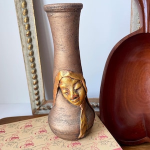 May include: A tall, slender vase with a textured, metallic bronze finish. The vase features a prominent, three-dimensional relief sculpture of a human face in a golden hue, positioned on the lower body, with closed eyes and a serene expression.
