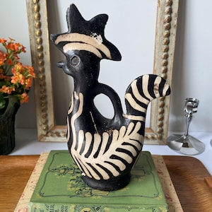 May include: A black ceramic bird-shaped vessel featuring a stylized crest and tail with cream-colored painted line details. The sculpture has a central circular handle opening and a smooth, matte finish, standing upright on a flat base.