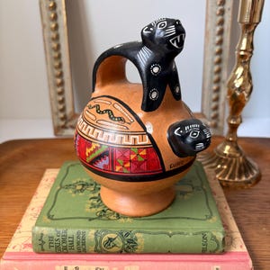 May include: A round terracotta vessel featuring a black jaguar handle and a smaller black jaguar head protruding from the side. The body is decorated with painted geometric patterns, a snake motif, and the word Cusco written in black ink.
