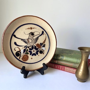 May include: A round ceramic plate with a speckled beige finish and a brown rim. The center features a hand-painted design of a stylized bird with a long beak, surrounded by dark blue floral motifs, brown circular accents, and a thin blue circle.