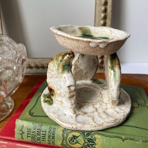 May include: A handmade ceramic oil burner with a speckled off-white glaze and green accents. The structure features a circular base, three curved upright supports, and a shallow bowl top, all crafted with a textured, organic, and uneven finish.