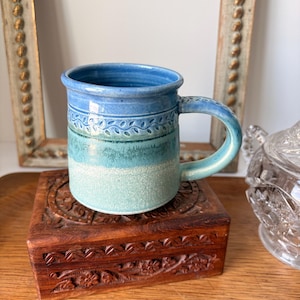 May include: A ceramic mug featuring a gradient of blue and teal colors. The upper section is a deep blue with a textured leaf pattern band, transitioning into a lighter teal and white speckled base. It has a curved handle and a smooth rim.