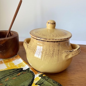 May include: A yellow ceramic sugar bowl with a matching lid and two side handles. The bowl features a distressed brown finish along the rim and lid edge. Beside it sits a small wooden bowl with a thin wooden stirring stick inside.