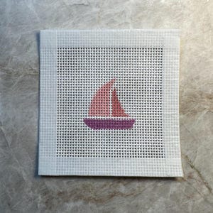May include: A square piece of white needlepoint canvas featuring a small, centered cross-stitch design of a sailboat. The boat has a purple hull and a pink sail, stitched directly onto the grid of the white fabric with taped edges.