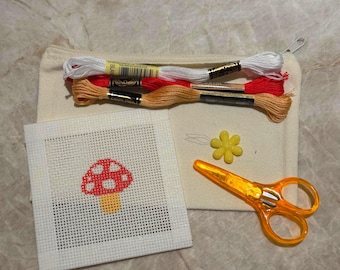 Puede incluir: Un kit de bordado que incluye un lienzo cuadrado blanco con un diseño de seta roja y blanca, tres madejas de hilo en blanco, rojo y beige, un imán para agujas amarillo con forma de flor y unas tijeras naranjas dentro de una bolsa de tela.